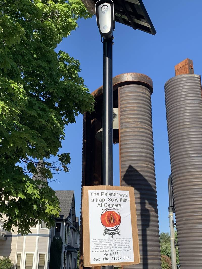 Flock camera on a pole with community flyer reading 'The Palantir was a trap. So is this AI Camera.' DeFlock.org